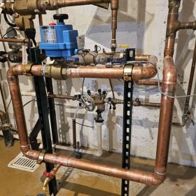 Copper pipes form a complex network against a concrete wall, featuring a blue valve and gauges. A yellow warning sign adds a cautionary tone.